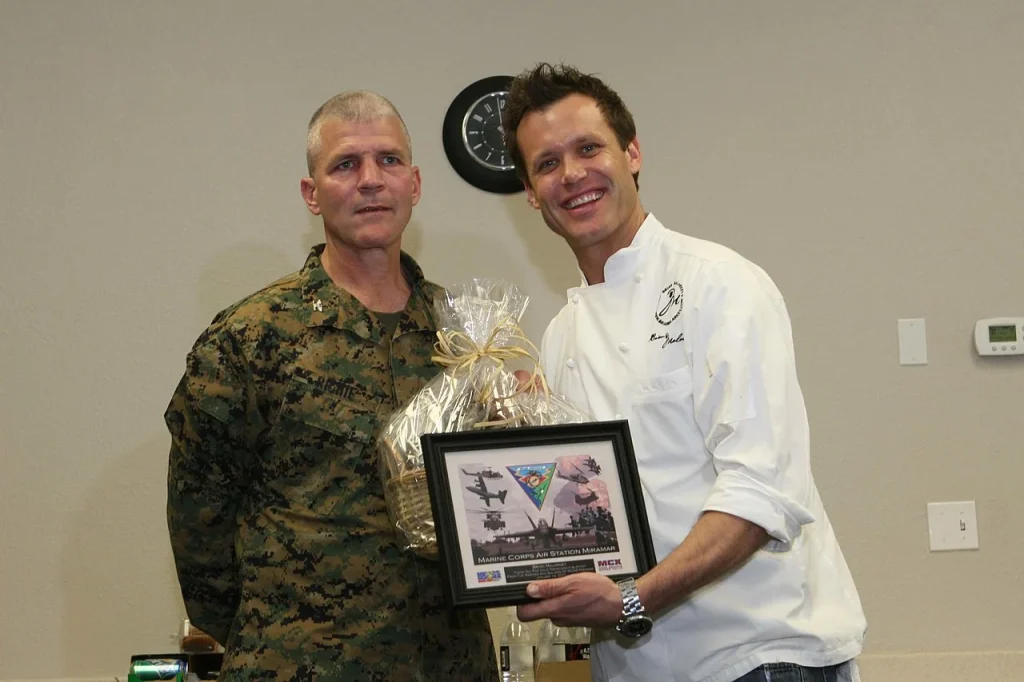 Chef Brian Malarkey in a white chef coat holding an award frame alongside a U.S. Marine Corps officer.