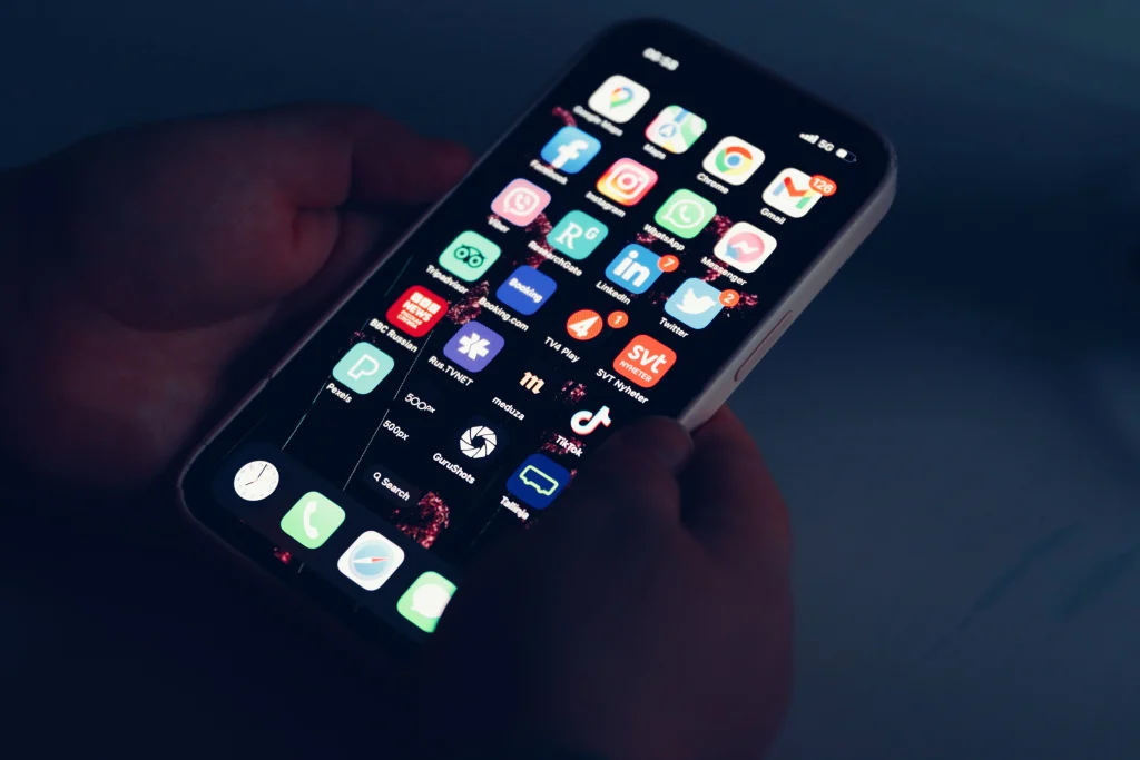 Person holding a smartphone in low light, displaying a home screen filled with various social media, news, and utility app icons.