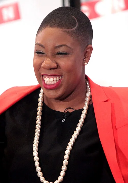 Symone Sanders smiling in a coral blazer and pearl necklace during her years as a CNN political analyst