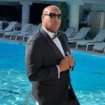 Mister Lewis Lux in a black tuxedo and sunglasses posing beside a luxury resort pool
